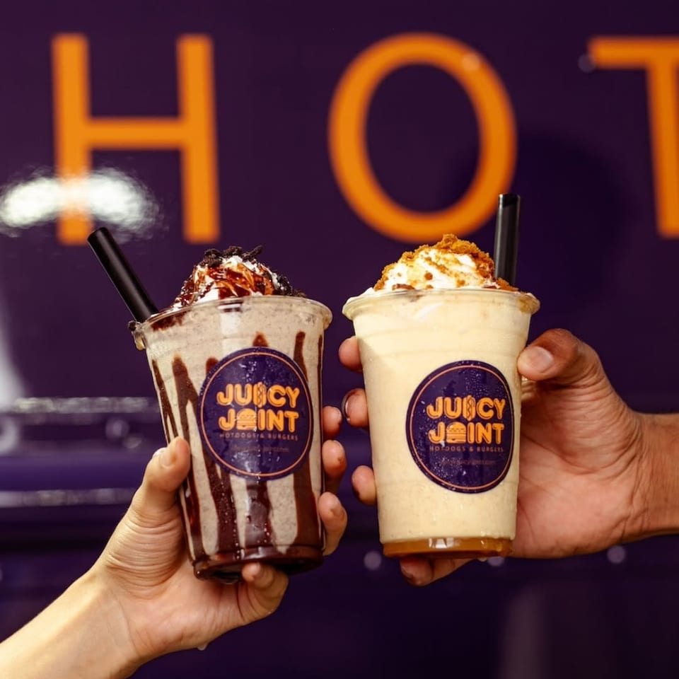 Two hands holding milkshakes from Juicy Joint, one chocolate and one vanilla, against a purple background.
