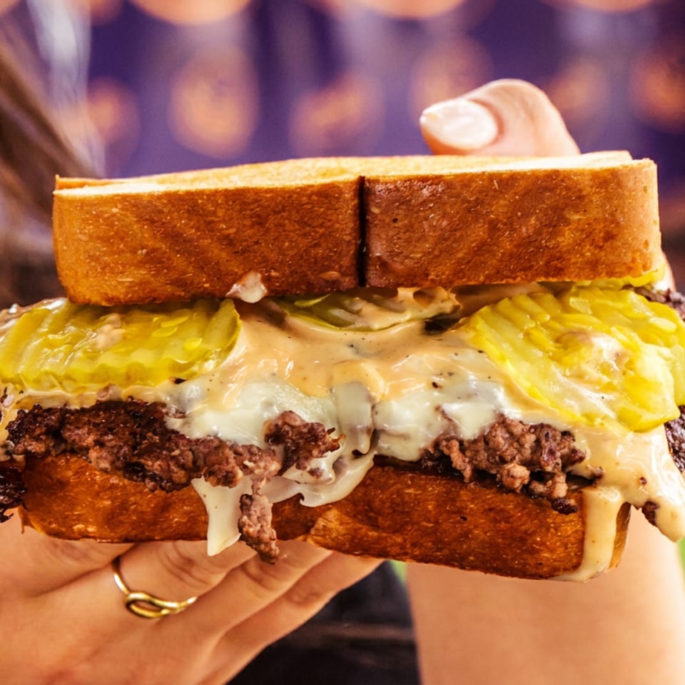 Hand holding a patty melt with melted cheese, pickles, and sauce on toasted bread.