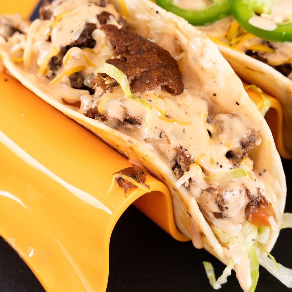 Two tacos filled with meat, cheese, lettuce, sauce, and jalapeños, resting in an orange holder.